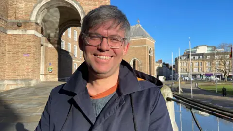 Councillor Barry Parsons is standing outside Bristol City Hall in a blue jacket, smiling at the camera. It is sunny and the sky is blue.