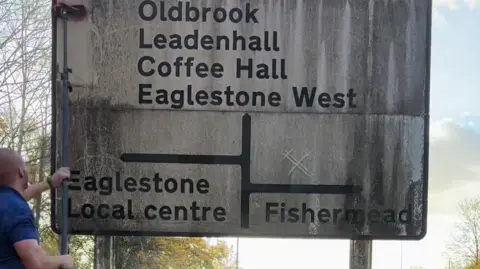 TikTok/The Ginger Cleaner A man cleaning a large white road sign with several places on it.