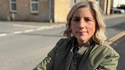 A head and shoulder shot of Jess on her mobility scooter in the street. She has short blonde hair and wears a khaki green jacket on top of a grey jumper. She looks at the camera with a neutral expression.