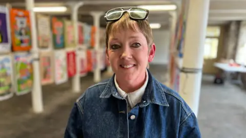 Sue Bardwell smiles at the camera. She has her glasses balanced on top of her head and is wearing a denim jacket. Blurred behind her is a colourful art exhibition.