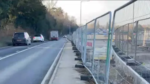 The A134 with metal fencing to the right, showing a housing estate being built, and cars driving along the road on the left. There are about four cars on the road, including one lorry. 