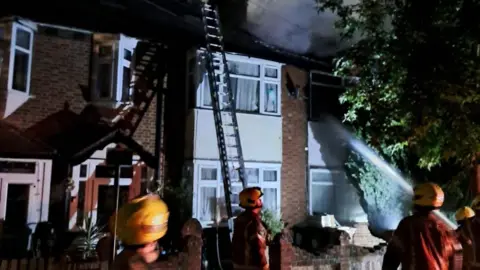 LFB Firefighters wearing orange jackets and helmets standing outside a terraced house, a ladder is leaning up against it and a hose and stream of water is being aimed up at the roof.