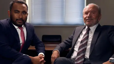 BBC Amol Rajan and Lord Sugar, sat down and wearing suits, looking into the camera