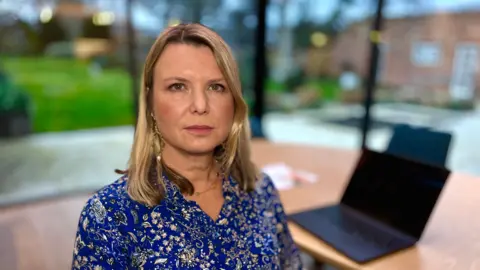 Martin Giles/BBC Dr Diana Hunter looks directly at the camera. She is in front of her laptop working from home. There are floor to ceiling windows all around her, in her study and you can see a grassed area through the windows. Her black laptop is placed on a large table. She is wearing a royal blue dress with silver flowers and has shoulder length blonde hair.