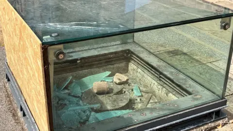 BBC/ Alec Blackman A glass display case with an object inside is boarded up on the left hand side. The glass panel has been smashed and the broken glass is covering the interior and the artefact inside,