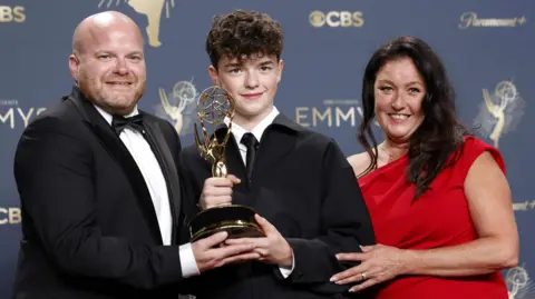 EPA Owen Cooper, who wears a black tie over a white and a black jacket, poses with the Emmy award, which shows a golden figure holding aloft a massive globe shape. His dad, who wears a black bow tie and suit, is on the left and mum, in a red evening gown, is on the right. They pose in front of the Emmy Awards backdrop at a photocall.
