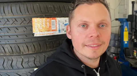 Joe Fisher, 35, has short brown hair swept across his head and has a slightly stubbled chin, he is wearing a black hoodie with the hood down, behind him are a stack of new tyres and equipment for changing tyres to the right.