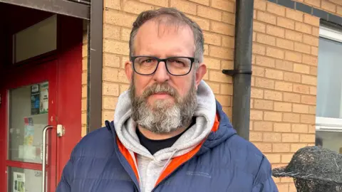 Andy Cammiss, standing in front of East Durham Veterans Trust in Seaham. He is dressed in a blue waterproof jacked with a grey hoody, is wearing black rimmed glasses and has a thick brown and grey beard and short brown hair.