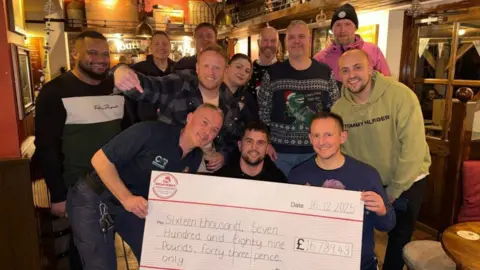 WMAS A gathering of people in a pub with a large cheque holding a large cheque. One man is pointing down towards the cheque. 