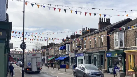 This image shows a high street lined with brick buildings: a mix of shops and businesses. A two-lane road runs through the center of the scene with colorful triangular bunting flags strung across the street. Strings of small overhead lights are also suspended above the road.
