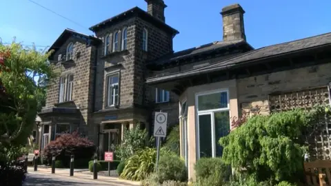 A stone building with trees around the driveway. It has a turret and two chimneys and there is a series of bollards and a road sign at the front. 