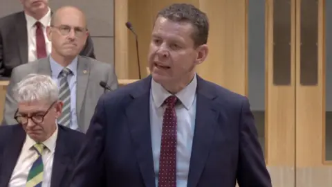 A man with a navy blue blazer, a light blue shirt and a red tie is stood up looking to the right. There are multiple men in the background as well as wooden doorway. 