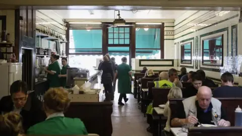 Getty Images People sitting at tables and eating in a cafe. Waitress wearing green dresses are standing and walking around.