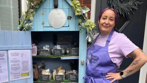 Dani wearing a purple apron and purple t-shirt standing next to the cake shed. The door is open and there are three shelves of cakes. The shed is blue and has a floral garland on the top