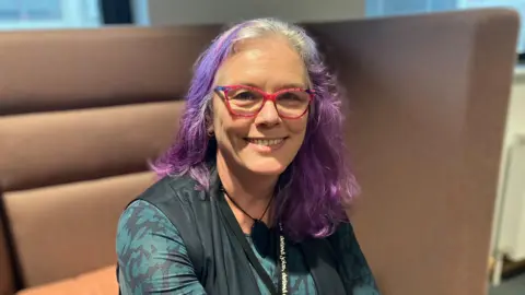 A woman with purple hair sitting on a sofa. She has bright pink, thick-rimmed glasses on and is smiling at the camera.