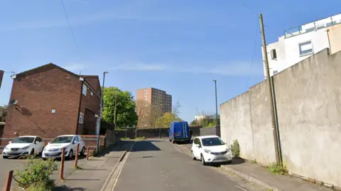 A Google street view of Sarah Street in Bristol, it is a short street with a small car park on the left and a tall wall on the right. There are council tower blocks in the background.