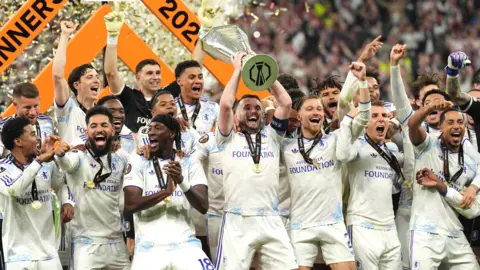 PA Media The Aston Villa team, made up of about 20 men, all wearing white uniforms with blue accents, are grouped together cheering. In the centre one of them is holding a large trophy above his head