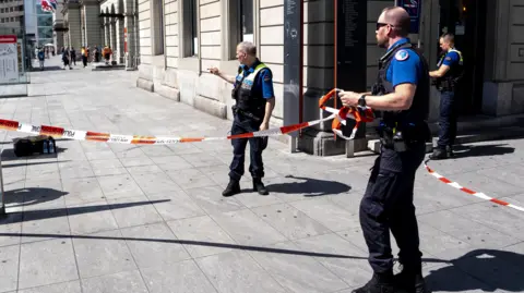 EPA Three police officers wearing black trousers, blue shirts and black and reflective police vests stand outside Winterthur train station. One officer wearing sunglasses is holding a roll of orange police tape.