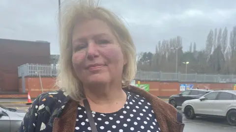 Pat Ward, wearing a spotty dress and a jacket and handbag, stands in the car park at Morrisons in Stirchley. There are parked cars behind her and the walls of the supermarket building, with trees in the background, and the sky is cloudy and grey.