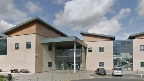 Google A large two storey building has an entrance off centre to the left which is made of glass and has two doors. There are disabled parking spaces in front of the building where two cars are parked.