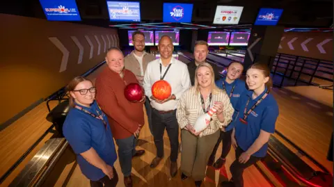 A group of people are standing on a lane in a bowling alley. Two of the men are holding bowling balls and a woman is holding a skittle.