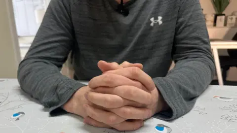 BBC A man, shown from the shoulders down, wears a grey long sleeved top and has his hands clasped on a table with a tablecloth with colourful robins on it. 