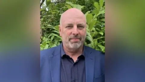 A man in a blue shirt and jacket standing outside in front of some greenery.