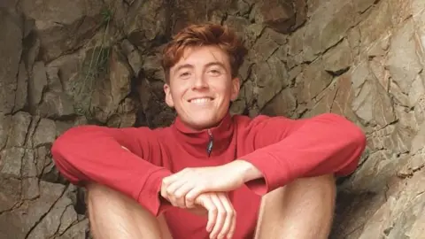 Tomos Sleep who has short light brown hair wearing a red jumper and shorts sat down. 