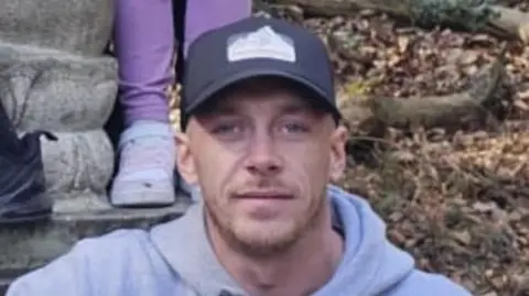 Dorset Police Joey Johnstone poses for a photograph outdoors. He wears a baseball cap and a light grey hoodie and has a short beard.