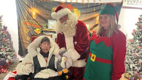 A young woman in a wheelchair has her photo taken with Father Christmas and a female elf. She has brown hair and wears a white puffer coat with a fur trim hood and a safety harness. 