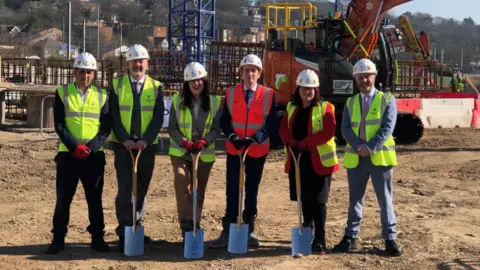 Rachel Hopkins, Labour MP for Luton South and South Bedfordshire, stands for a photo with other local dignitaries on the site of The Stage flagship regeneration scheme. They are wearing hard hats and fluorescent jackets with construction work happening in the background.