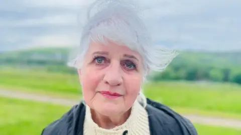 Ishbel Shand, a woman with grey hair and wearing a cream knitted top and a dark jacket, with a grassy landscape behind her.