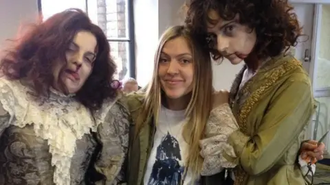 Anna Tennant (middle) pictured on the set of a film called Limehouse Golum, with straight blonde hair and wearing a while t-shirt and a necklace. She is surrounded by two other women wearing period costumes and makeup where they look like zombies