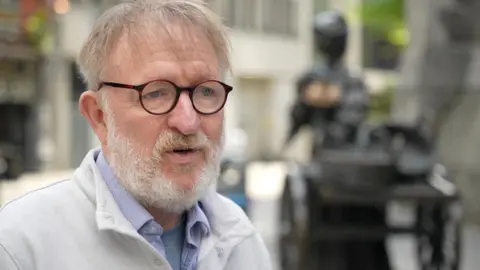 A white haired man with a beard, black rimmed glasses and a grey jumper