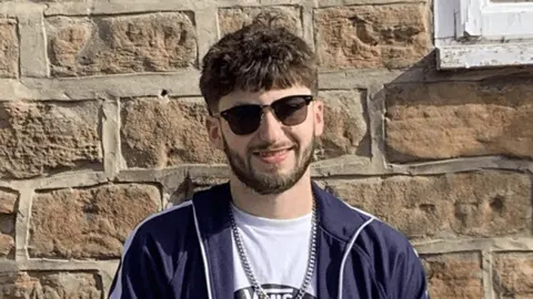 James Bosworth wearing a blue jacket, white Vans T-shirt with a silver chain and sunglasses in front of a stone wall