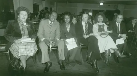 Fumi Stephenson Dr and Mrs Stephenson (second and third left) sit with Paul Dankworth and Cleo Laine (to their right). They are sat in what appears to be a large hall, as though they are about to watch a performance, and are all dressed smartly. 