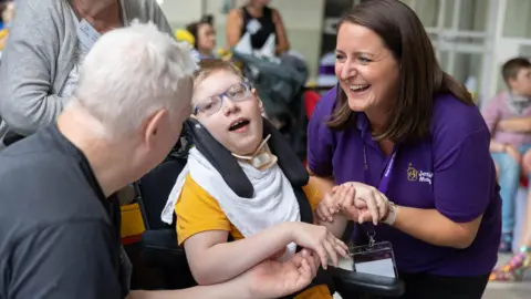 Jessie May A young boy in a wheelchair holding hands with a nurse from Jessie May, who is smiling and wearing a purple t-shirt