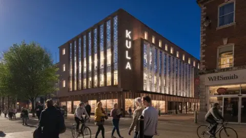 The outside of a hotel, which has the words 'Kula' written on it, showing the lights on at night. There are also pedestrians and cyclists passing by outside. 