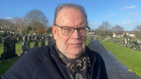 Historic Tom Hartley. A man wear square framed glasses, a scarf black and yellow and a jacket of the navy zip. Stay on a walk in the middle of a cemetery.