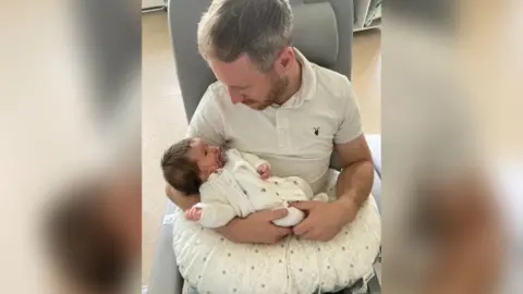 Pete Grimwade pictured sitting down holding his baby daughter, Isabella, in his arms, looking down at her while she looks up at him