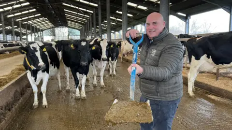 BBC A man in a padded jacket holding a blue spade full of manure towards the camera. He is standing in a cow shed and there are numerous black and white cows behind him.