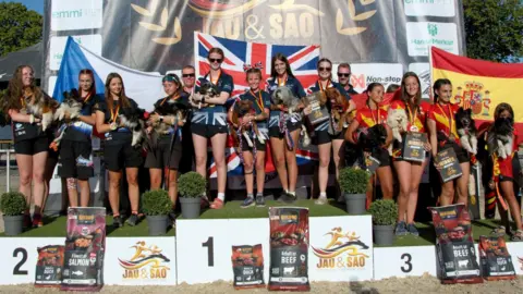 Gavin A large group of girls and women, standing on three podiums all holding dogs. Flags and a large banner are behind them. 