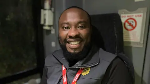 Pepu Kayinari smiles wearing a red lanyard and a grey gilet with the yellow Bee Network branded bee symbol. 