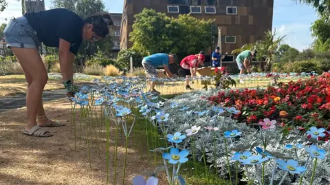 A woman in denim shorts and t-shirt crouches as she plants blue and purple metal forget-me-not flowers in grass next to a bed of white and red flowers. Others can be seen in the background doing the same. Houses are in the background.
