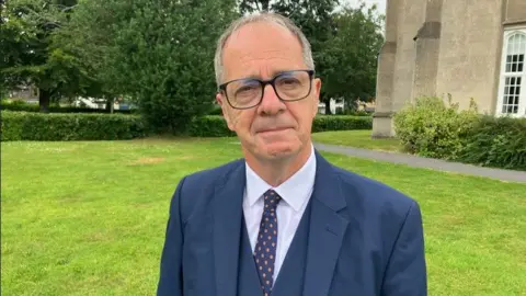 Emlyn Dole is smiling as he stands in front of the campus building. He has short grey hair and is wearing glasses, a light-coloured shirt, a blue tie with an orange dotted pattern and a three-piece suit.