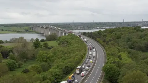 Vehicles queue on the A14 heading toward the Orwell Bridge. The River Orwell can be seen under the bridge and trees line either side of the carriageway leading up to this.