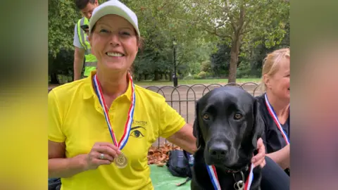 Irena Valchera A woman wearing a bright yellow shirt with an embroidered logo and a medal on a red‑white‑blue ribbon around their neck. She is holding a black labrador wearing a matching medal. Another person, also wearing a medal, is seated to the side. The group are in a park with trees, a fence, and grass in the background.