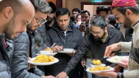 A group of people gather around a buffet-style food table, holding plates of rice and other dishes. Several individuals are serving themselves while others wait nearby in a crowded indoor setting.