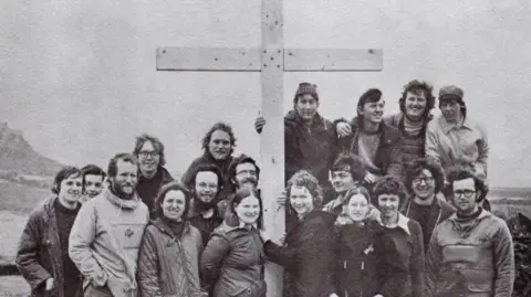 A black-and-white photograph of 19 young people standing around a wooden cross, four of them appear to be standing on something as they are higher than the others. To the right you can see the outline of Holy Island 