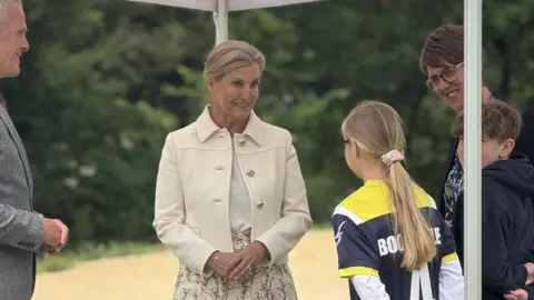 Blonde haired woman wearing a smart cream jacket and a floral maxi skirt talking to two children. One child has a  blonde ponytail and is wearing a yellow and black school p.e kit. And the brunette boy is wearing a black hoodie. The man is standing in the corner of the shot wearing a grey suit. 